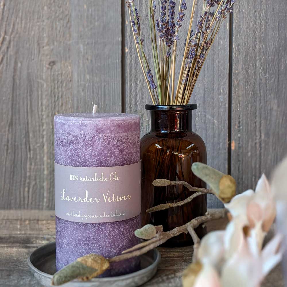 Lila Duftkerze mit der Aufschrift „Lavender Vetiver – 100 % natürliche Öle, von Hand gegossen in der Schweiz“. Daneben steht eine braune Glasvase mit getrocknetem Lavendel vor Holz-Hintergrund.