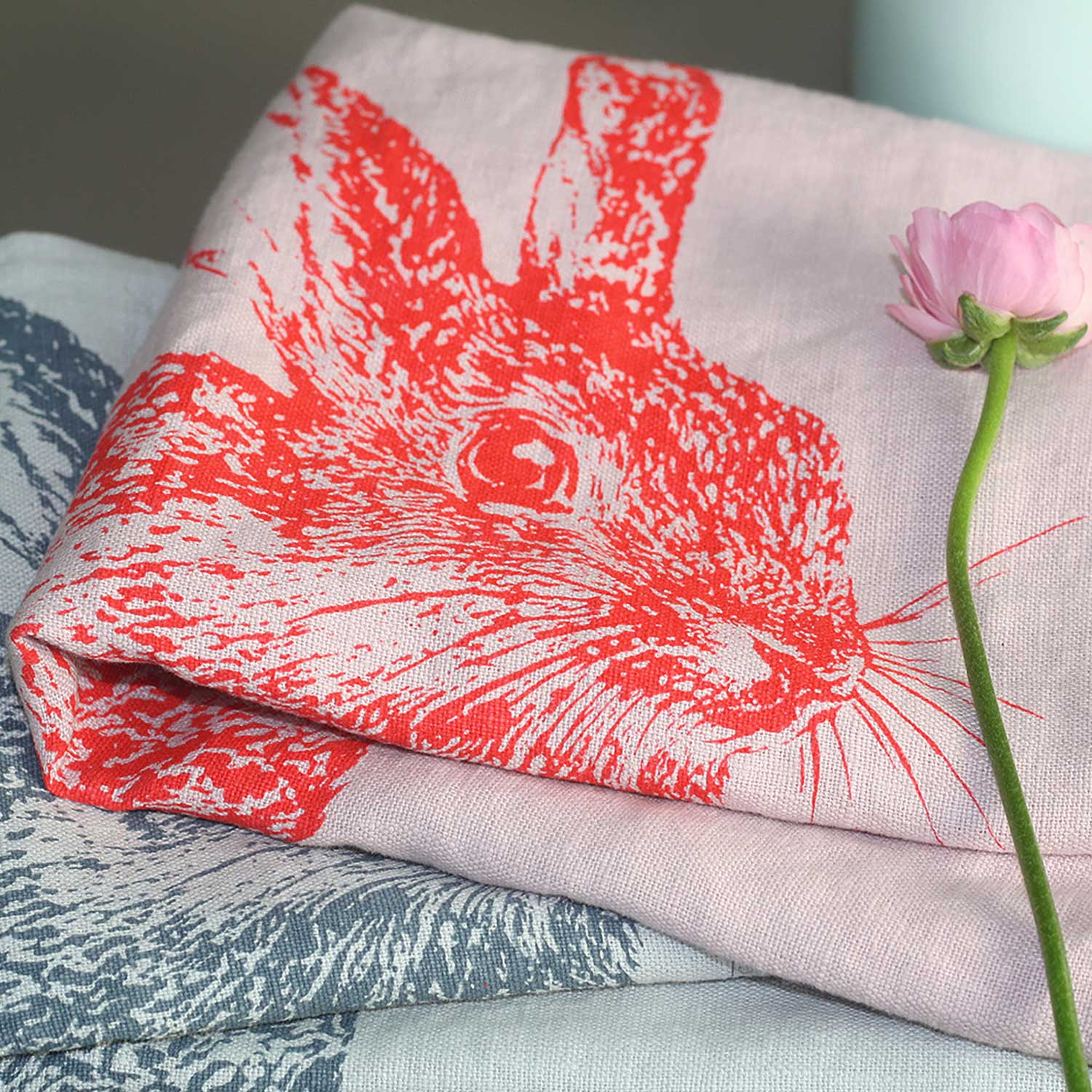 Handbedrucktes Frohstoff Geschirrtuch mit großem Kaninchenmotiv in Korallrot auf Leinen und Halbleinen, dekorativ inszeniert mit Blüte und pastelligen Naturtönen.