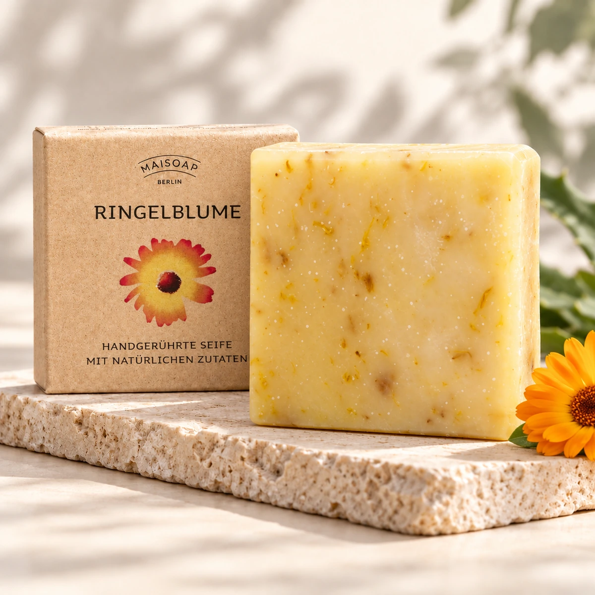 Maisoap Ringelblumenseife mit Blütenstückchen, Verpackung und orangefarbener Blüte auf Steinplatte