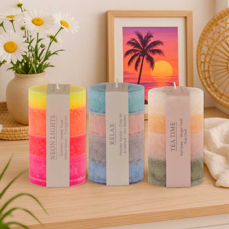 Drei mehrfarbige Duftkerzen mit Etiketten „Neon Lights“, „Relax“ und „Tea Time“ stehen auf einem hellen Holztisch. Im Hintergrund stehen Gänseblümchen in einer Vase und ein Bild mit Palmen und Sonnenuntergang.