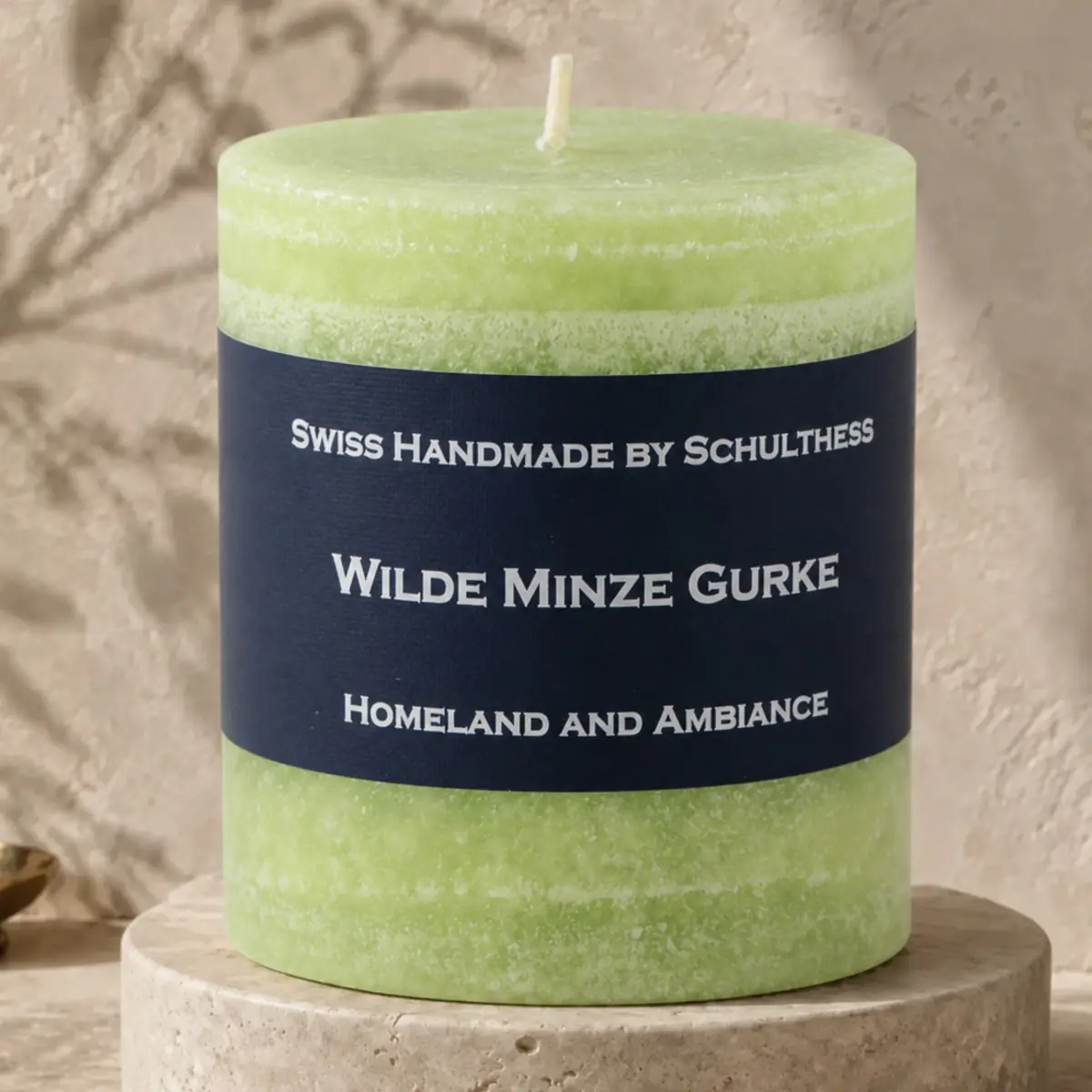 Hellgrüne Schulthess Duftkerze Wilde Minze Gurke auf einem runden Travertinsockel vor einer hellen, strukturierten Wand.
