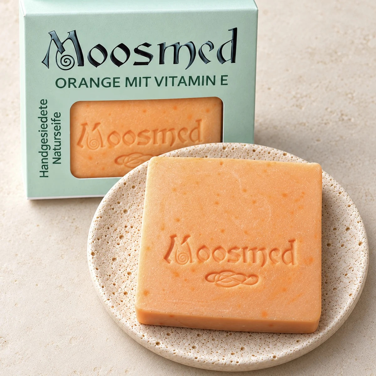 Orangefarbene Moosmed-Seife mit Vitamin E auf einem runden Travertinteller, dahinter die mintgrüne Verpackung.