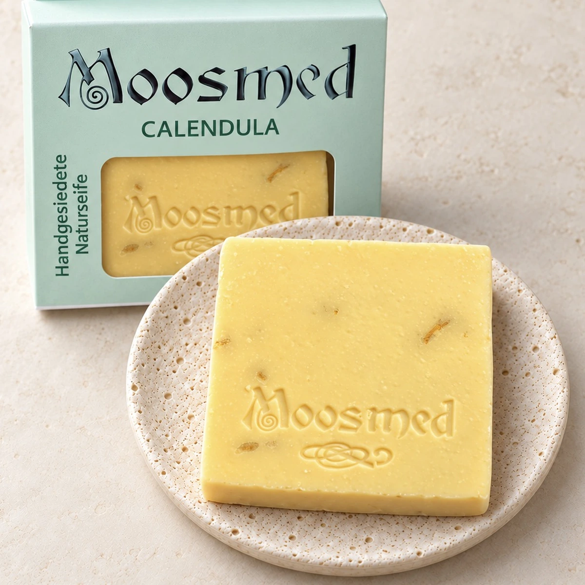Goldgelbe Moosmed-Calendula-Seife mit sichtbaren Blütenstückchen auf einem runden Travertinteller, dahinter die mintgrüne Verpackung.