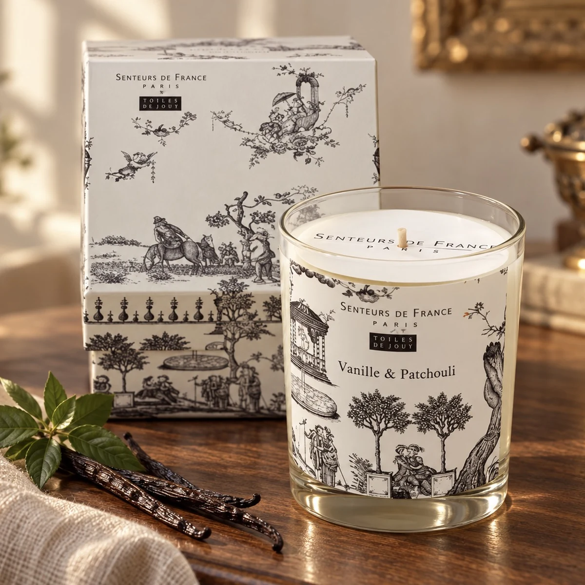 Duftkerze Vanille & Patchouli von Senteurs de France mit heller Toile-de-Jouy-Geschenkbox und Vanilleschoten auf Holz.