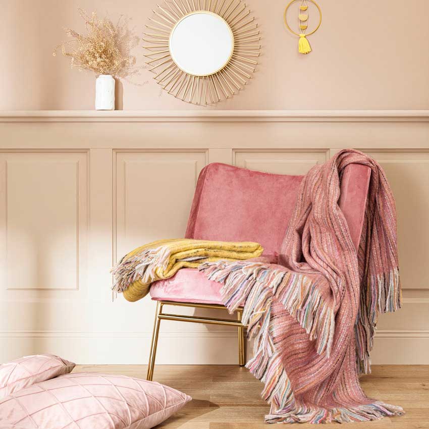 Magma Plaid-Serie „Ayana“ in sanften Pastellfarben – kuschelige Decke mit Fransen und passendem Kissen in Rosa und Gelb, stilvoll arrangiert auf Samtstuhl.
