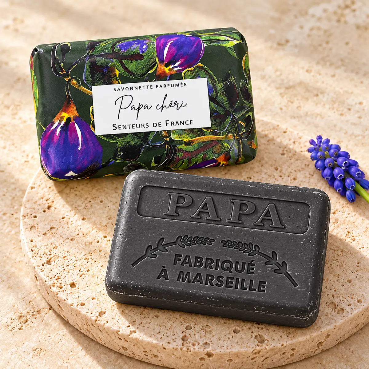Dunkle Papa-chéri-Seife mit Papa-Prägung auf Travertin, dahinter die grüne Blumenverpackung und eine violette Blütenrispe.