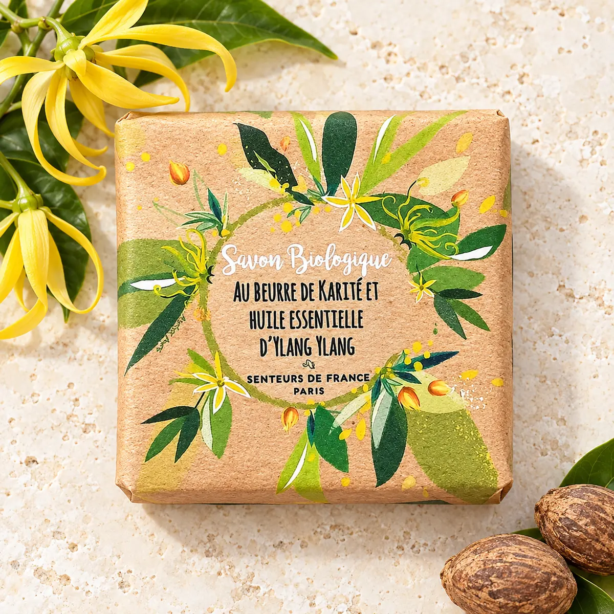 Verpackte Senteurs de France Bioseife mit Sheabutter und Ylang-Ylang, dekoriert mit gelben Blüten und Nüssen auf Travertin.