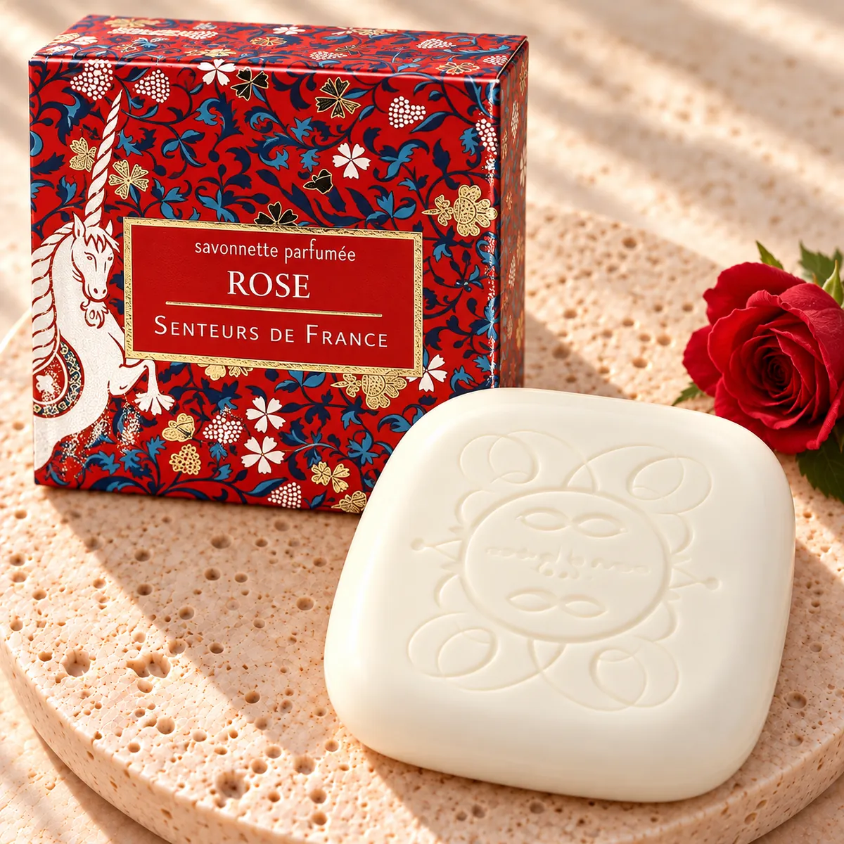 Cremefarbene Rosenseife mit Sonnenprägung auf Travertin, mit roter Einhorn-Verpackung von Senteurs de France und einer roten Rose.