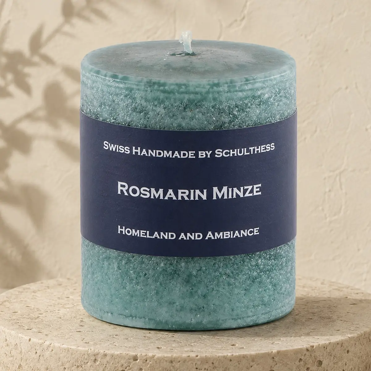 Türkisgrüne Schulthess Duftkerze Rosmarin Minze auf einem runden Travertinsockel vor einer hellen, strukturierten Wand.