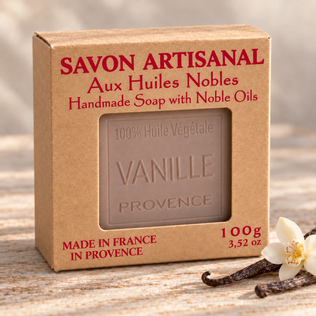 Maître Savonitto Seife Vanille in vollständig sichtbarer Kraftkartonverpackung mit Sichtfenster, neben Vanilleschoten und einer hellen Blüte auf hellem Holz.