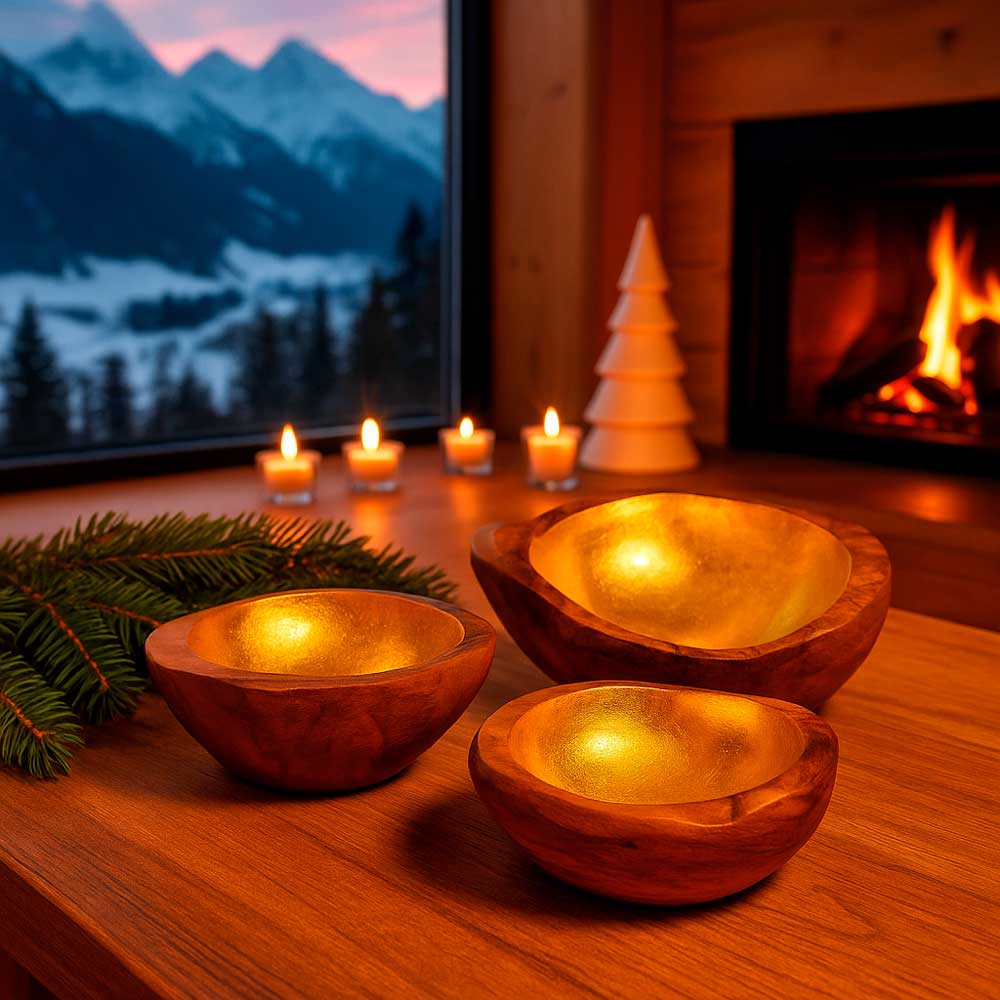 Drei Teakholzschalen mit goldfarbenem Inlay auf einem Holztisch vor einem Kaminfeuer. Im Hintergrund brennen Kerzen, und durch das Panoramafenster ist eine verschneite Berglandschaft zu sehen.
