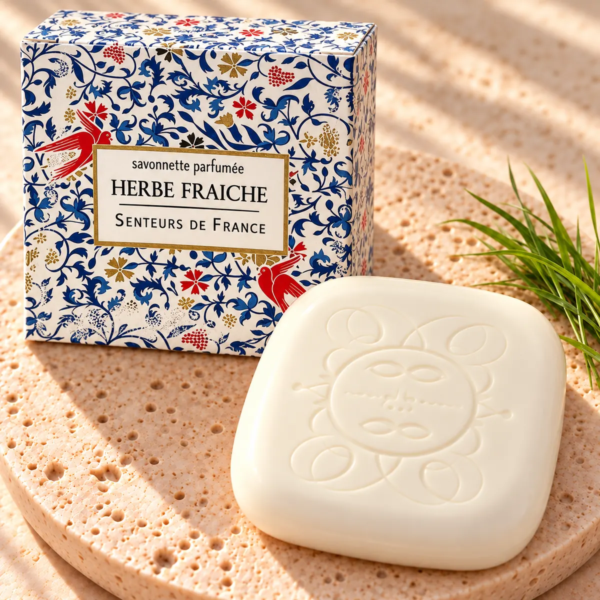 Cremefarbene Herbe-Fraîche-Seife mit Sonnenprägung auf Travertin, mit blau-weiß illustrierter Verpackung, roten Vogelmotiven und frischen Grashalmen.