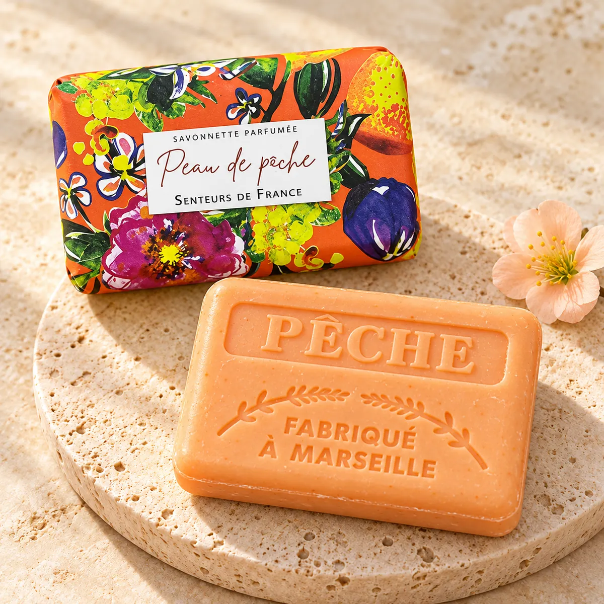 Pfirsichfarbene Peau-de-pêche-Seife auf einem Travertinteller mit orangefarbener Blumenverpackung und heller Blüte.