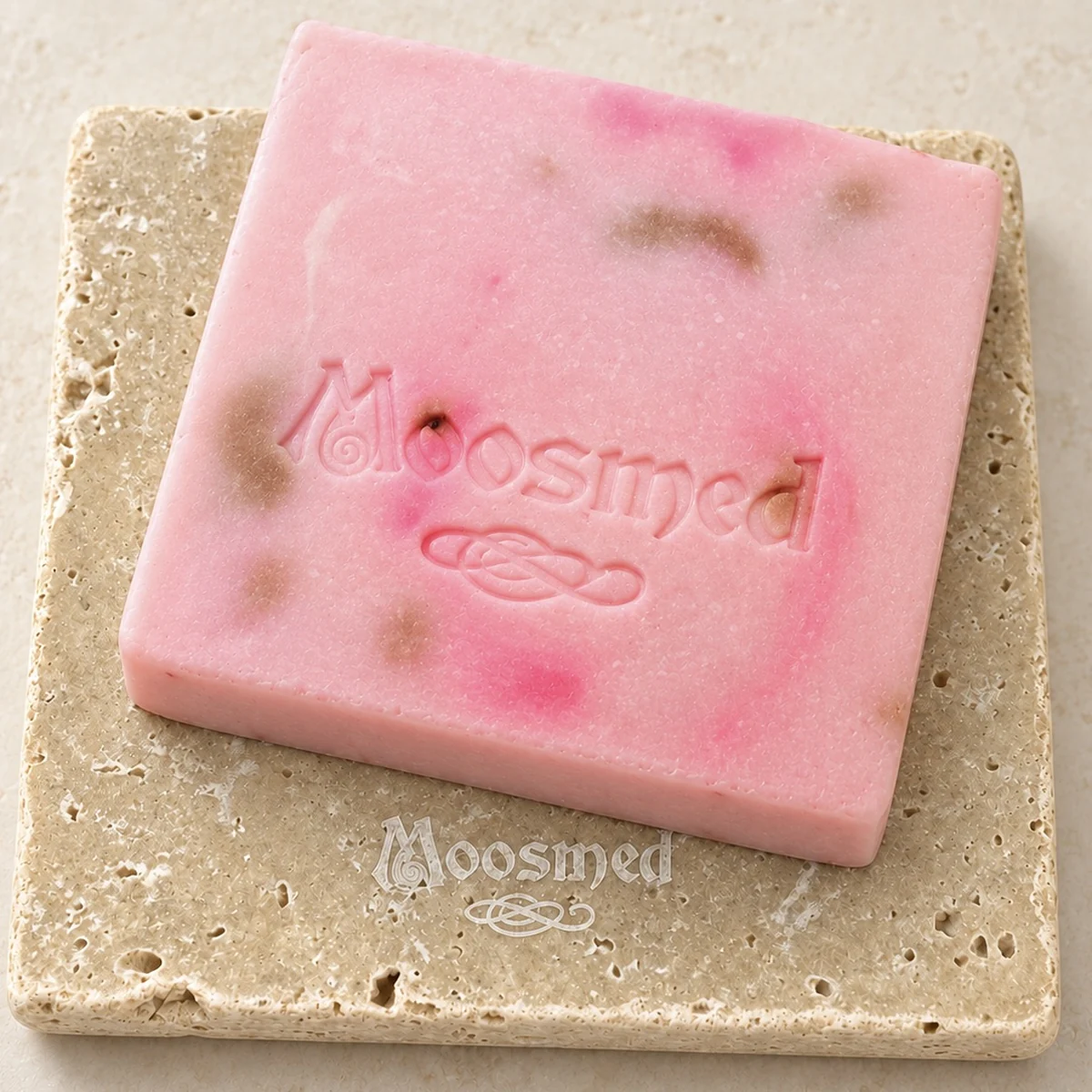 Rosafarbene Moosmed-English-Rose-Seife mit pinken und bräunlichen Akzenten und geprägtem Logo auf einem quadratischen Travertinteller.
