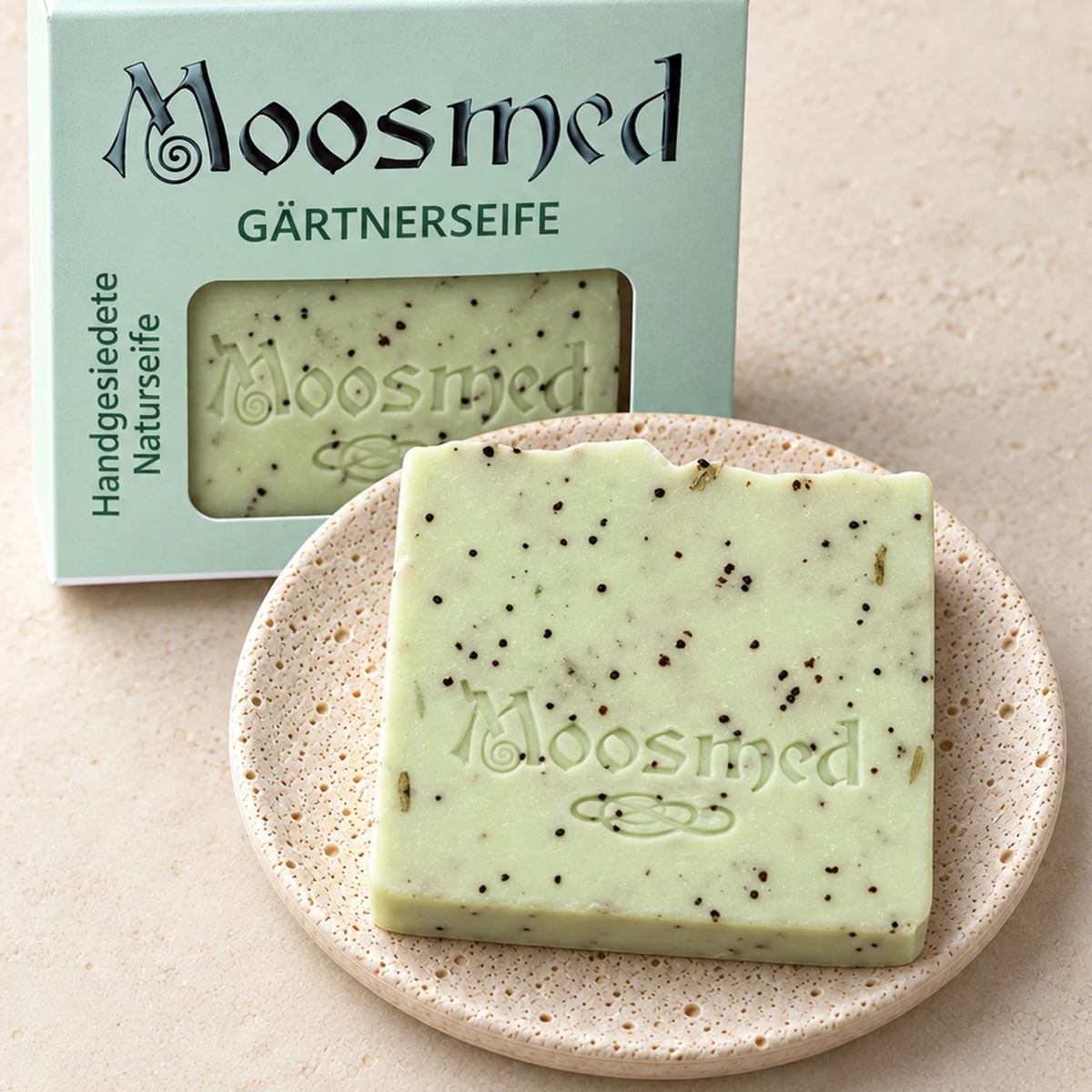 Hellgrüne Moosmed-Gärtnerseife mit dunklen Körnchen und Pflanzenstückchen auf einem runden Travertinteller, dahinter die mintgrüne Verpackung.
