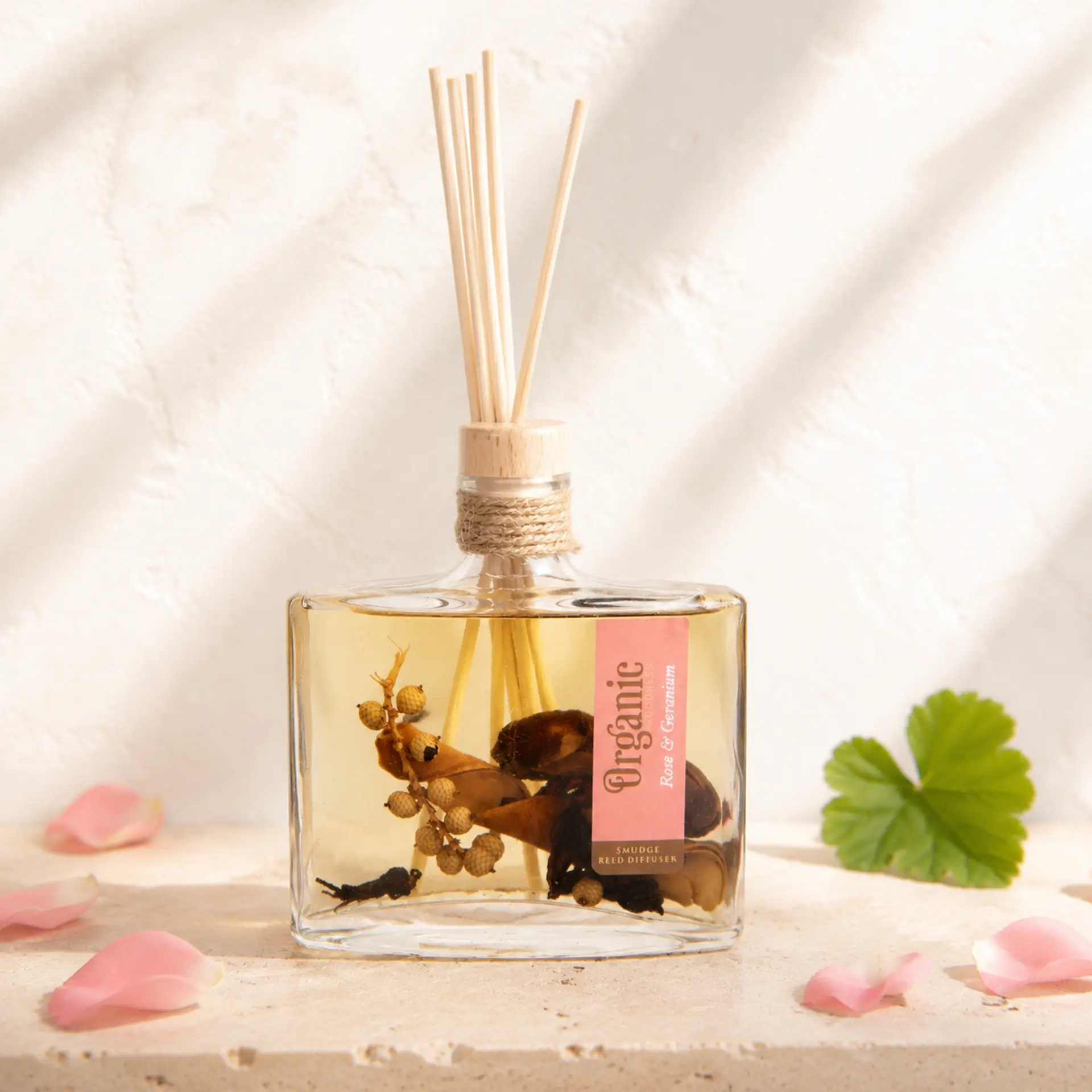 Organic Goodness Reed Diffuser Rose & Geranium mit Rosenblüten und Geranienblatt auf hellem Travertin.