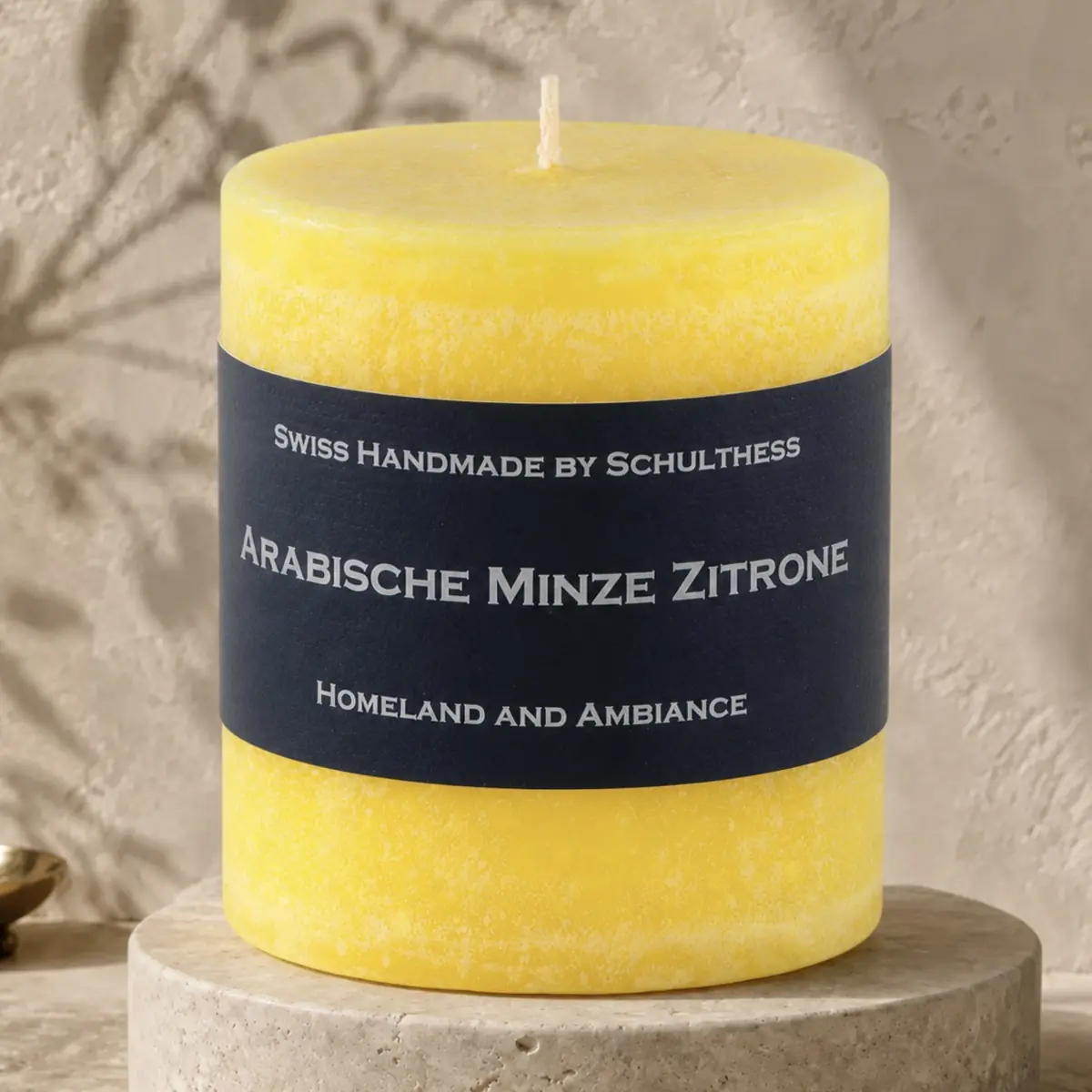 Gelbe Schulthess Duftkerze Arabische Minze Zitrone auf einem runden Travertinsockel vor heller, strukturierter Wand.
