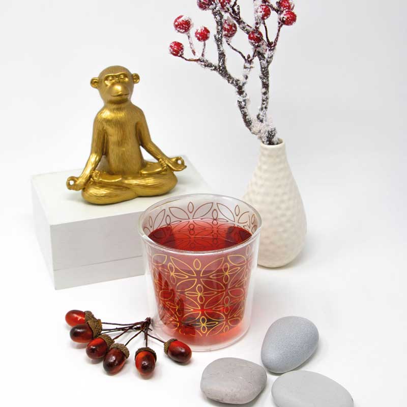 Stilvolle Szene mit rotem PPD-Teeglas im goldenen Ornamentdesign, daneben ein goldener Buddha-Affe, weiße Vase mit Beerenzweig, glatte Steine und winterliche Dekoration in sanften Farben.