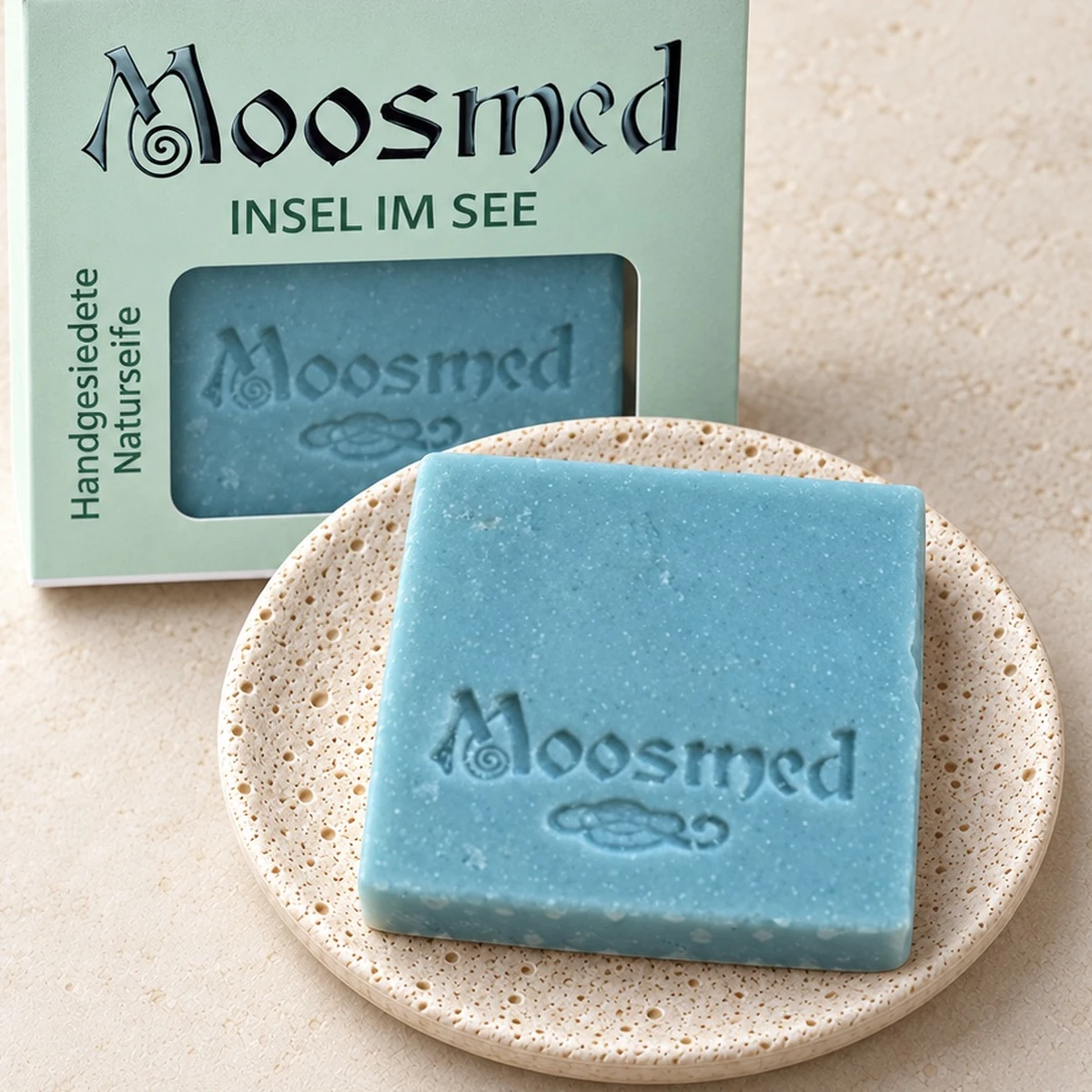 Türkisblaue Moosmed-Seife „Insel im See“ auf einem runden Travertinteller, dahinter die mintgrüne Verpackung.