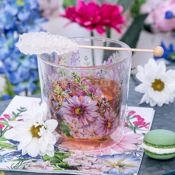 PPD Teeglas mit floralen Blütenmotiven in Rosa und Violett, arrangiert auf passender Serviette mit weißen Blumen und grünem Macaron – stilvolle Tischkultur für Frühling und Sommer.