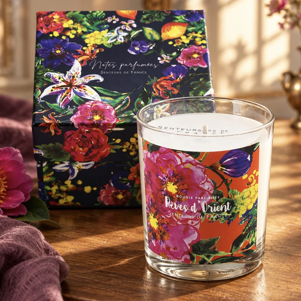 Duftkerze Rêves d’Orient von Senteurs de France mit floraler Geschenkbox auf dunklem Holz, neben Blüte und pflaumenfarbenem Leinen.