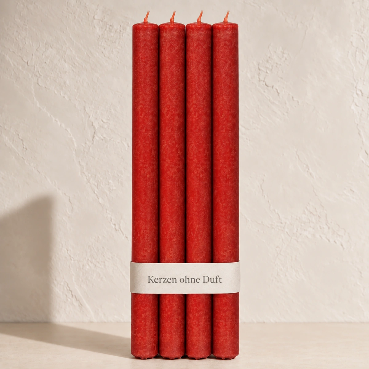 Vier Schulthess Stabkerzen in Tomatenrot mit beiger Banderole „Kerzen ohne Duft“ vor einer hellen, warmen Wand.