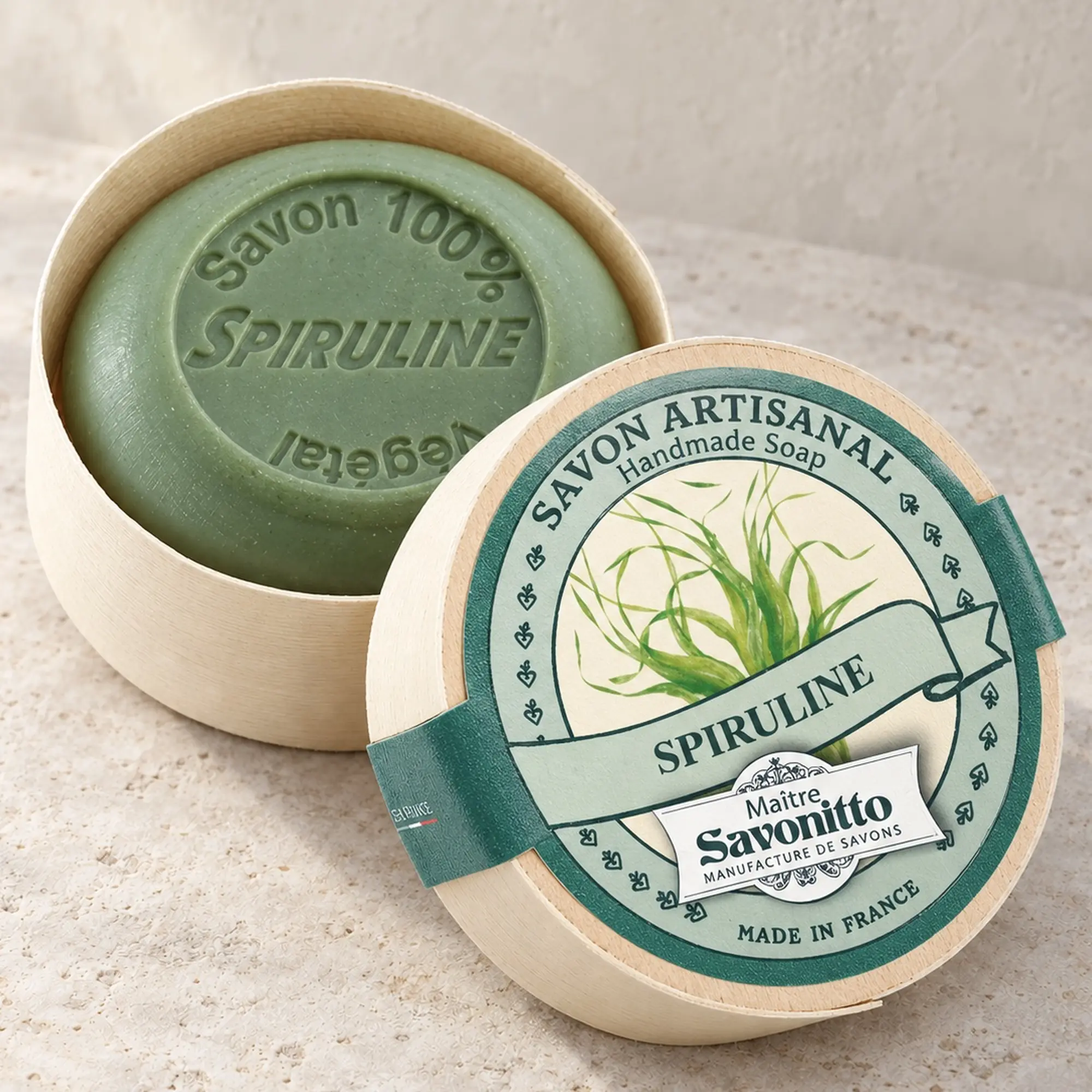 Maître Savonitto Spirulina in offener runder Holzschachtel mit angelehntem Etikettdeckel auf heller Steinfläche.