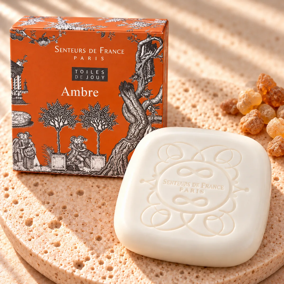 Cremefarbene Ambre-Seife von Senteurs de France mit ornamentaler Prägung auf Travertin, mit orangefarbener Toile-de-Jouy-Schachtel und rosa Beeren.