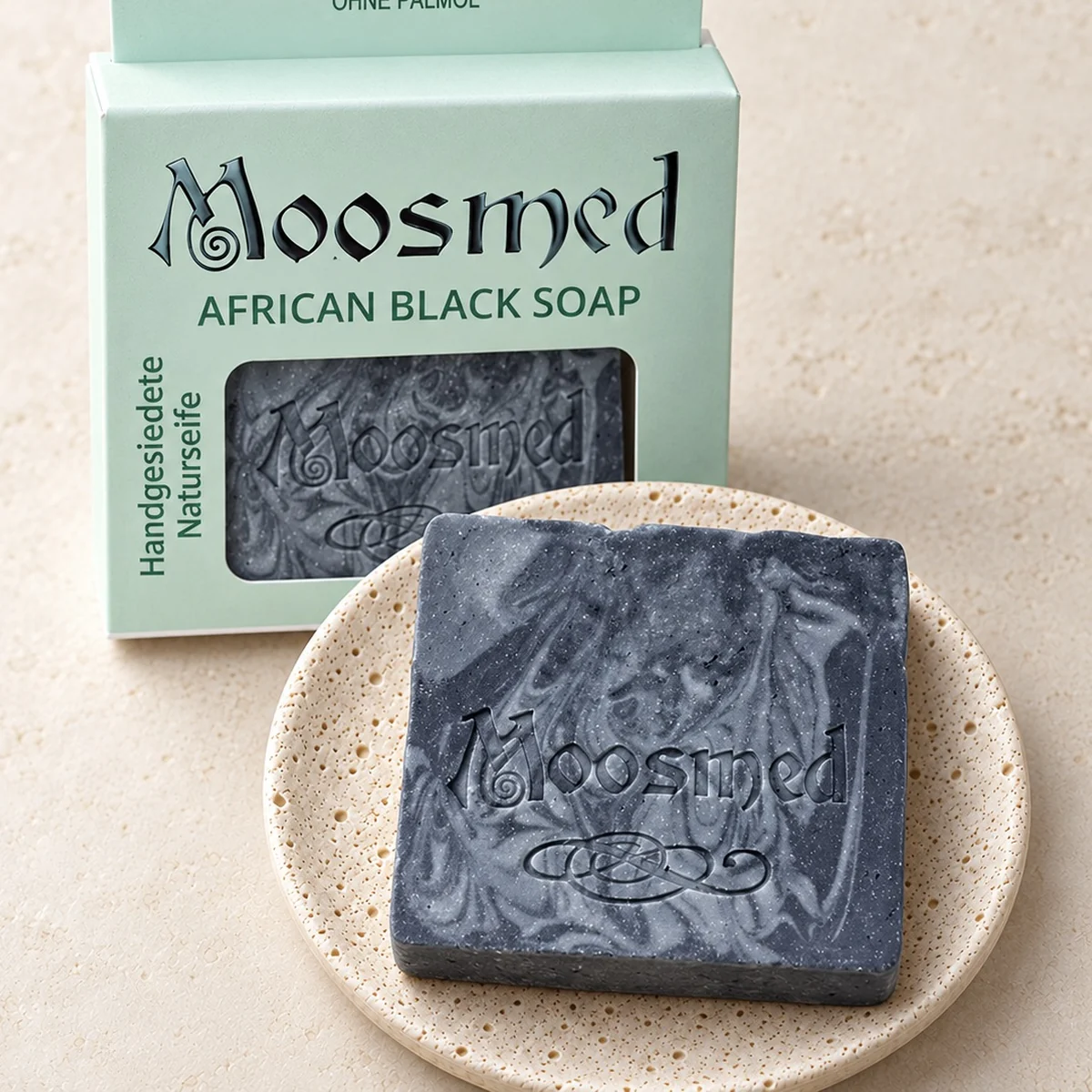 Dunkelgraue Moosmed African Black Soap mit heller Marmorierung auf einem runden Travertinteller, dahinter die mintgrüne Verpackung.