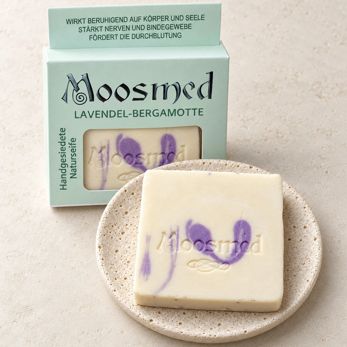 Cremefarbene Moosmed-Lavendel-Bergamotte-Seife mit violetter Marmorierung auf einem runden Travertinteller, dahinter die mintgrüne Verpackung.