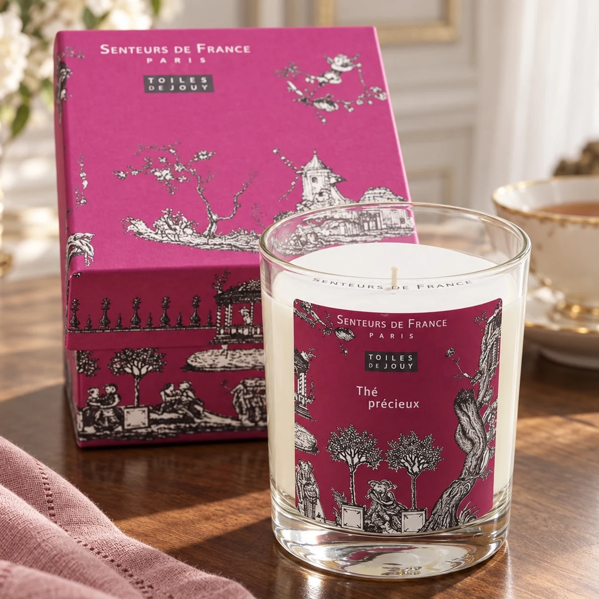 Duftkerze Thé Précieux von Senteurs de France mit pinker Toile-de-Jouy-Geschenkbox, roséfarbenem Leinen und Teetasse auf dunklem Holz.