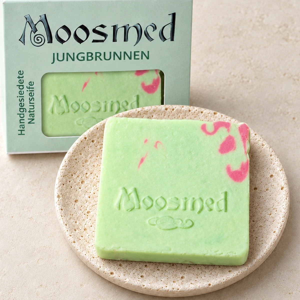 Hellgrüne Moosmed-Jungbrunnen-Seife mit pinker Marmorierung auf einem runden Travertinteller, dahinter die mintgrüne Verpackung.