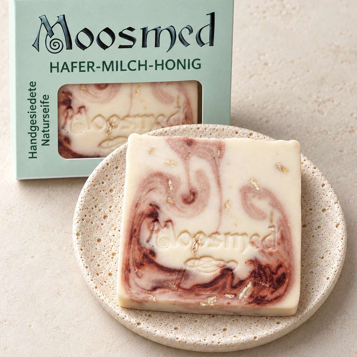 Cremefarbene Moosmed-Hafer-Milch-Honig-Seife mit rötlicher Marmorierung auf einem runden Travertinteller, dahinter die mintgrüne Verpackung.