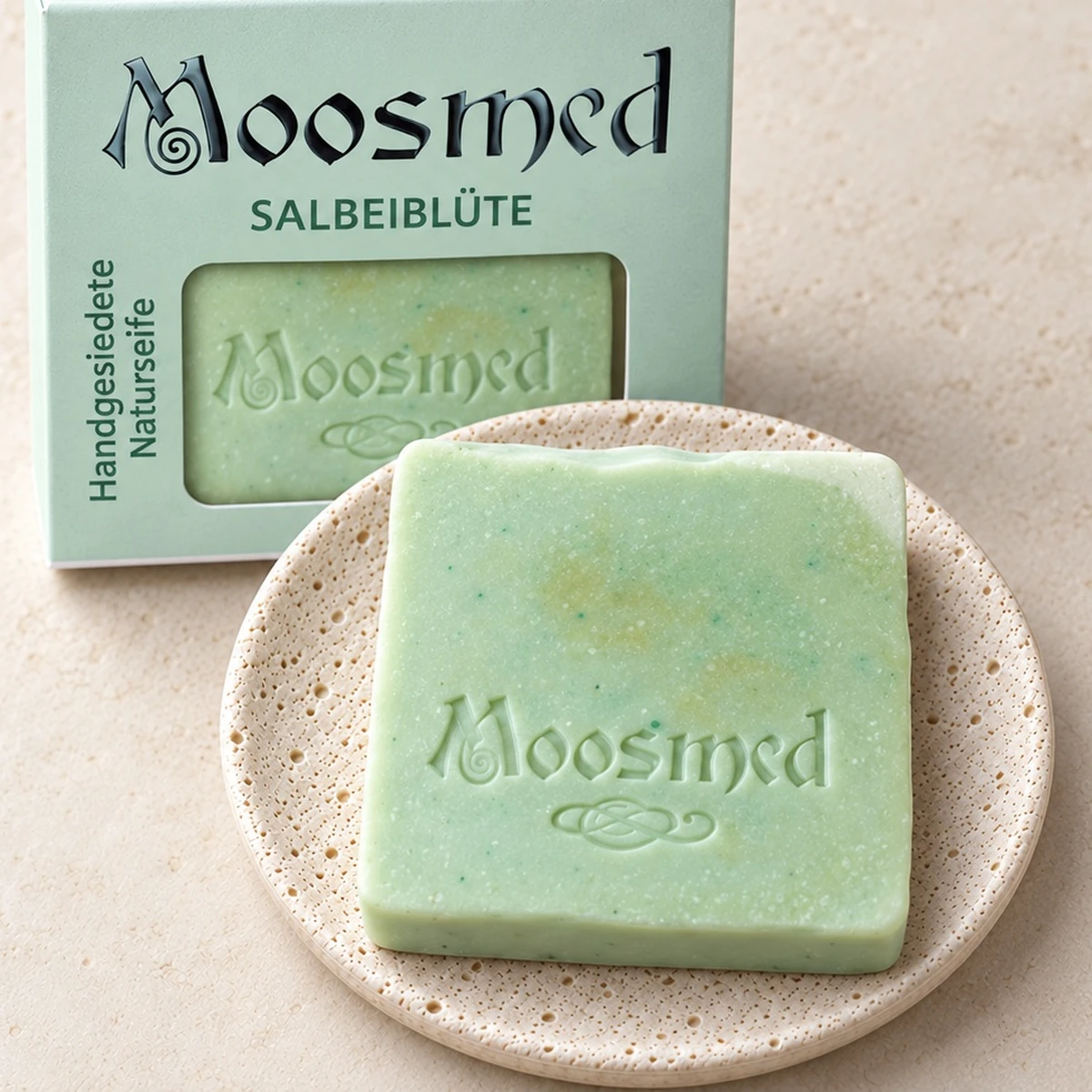 Hellgrüne Moosmed-Salbeiblüte-Seife mit feinen Sprenkeln auf einem runden Travertinteller, dahinter die mintgrüne Verpackung.