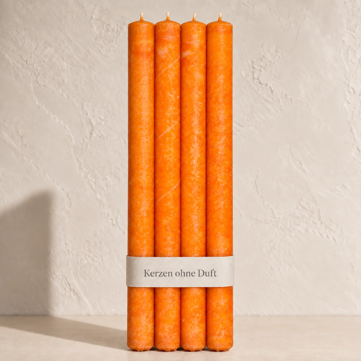 Vier Schulthess Stabkerzen in Mandarine mit beiger Banderole „Kerzen ohne Duft“ vor einer hellen, warmen Wand.