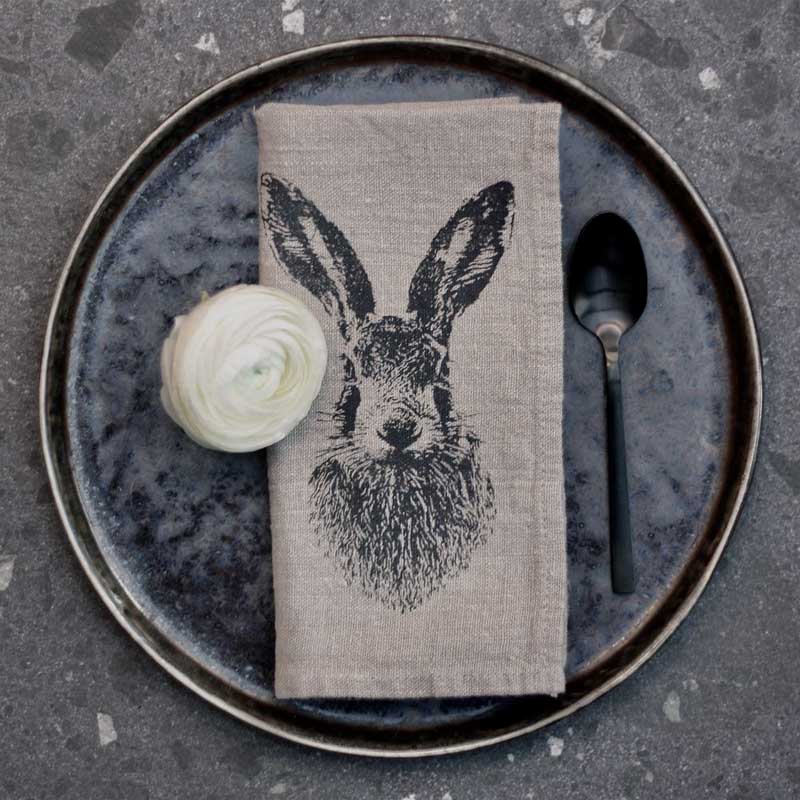 Leinenserviette von Frohstoff mit Feldhasen-Motiv in Anthrazit, stilvoll auf dunklem Keramikteller mit Blüte und Besteck arrangiert – natürliche Tischdeko.
