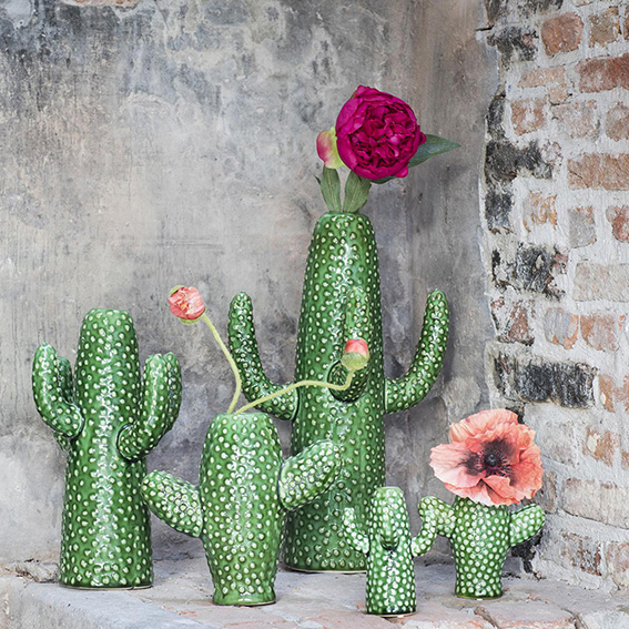 Dekorative grüne Vasen in Kaktusform mit weißen Punkten, arrangiert vor einer Beton- und Ziegelwand, einige mit bunten Blüten in Rosa und Orange geschmückt.