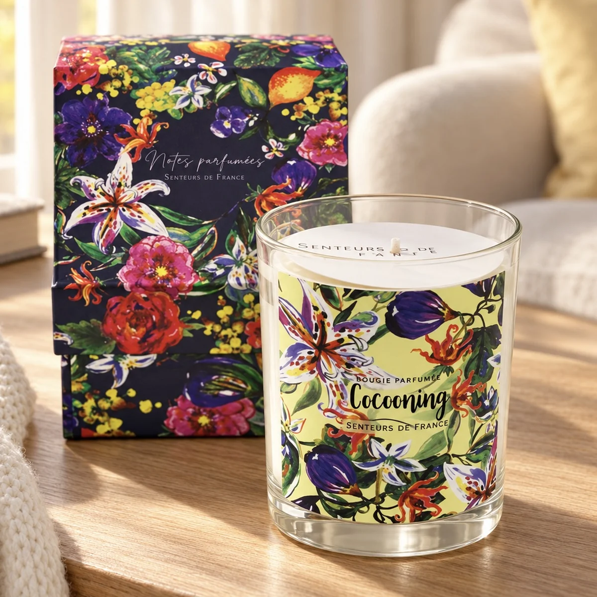 Duftkerze Cocooning von Senteurs de France mit floraler Geschenkbox auf hellem Holz neben einer cremefarbenen Strickdecke.