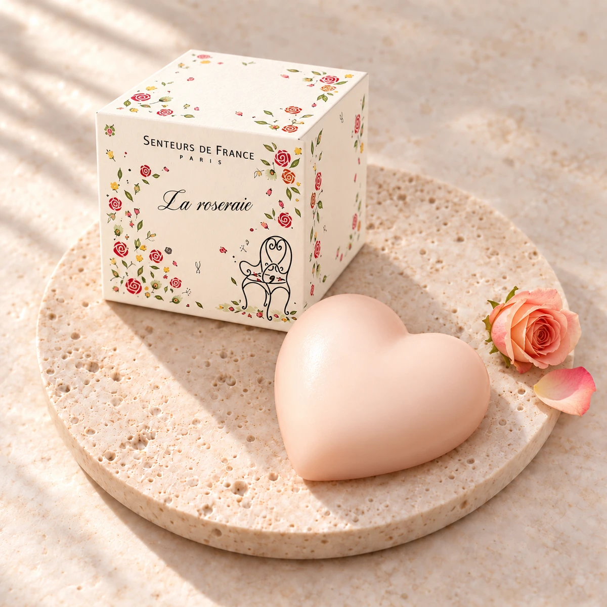Zartrosa Herzseife La Roseraie von Senteurs de France auf einem Travertinteller mit rosengeschmückter Würfelschachtel, einer Rose und einem Rosenblatt.