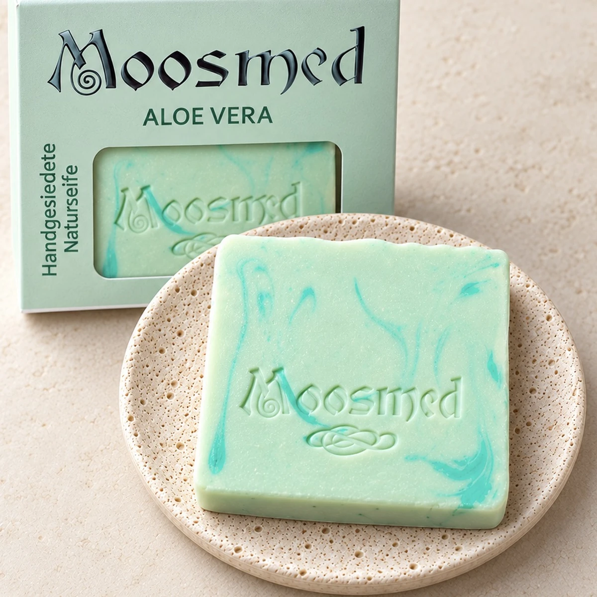 Mintgrüne Moosmed-Aloe-Vera-Seife mit türkisfarbener Marmorierung auf einem runden Travertinteller, dahinter die mintgrüne Verpackung.