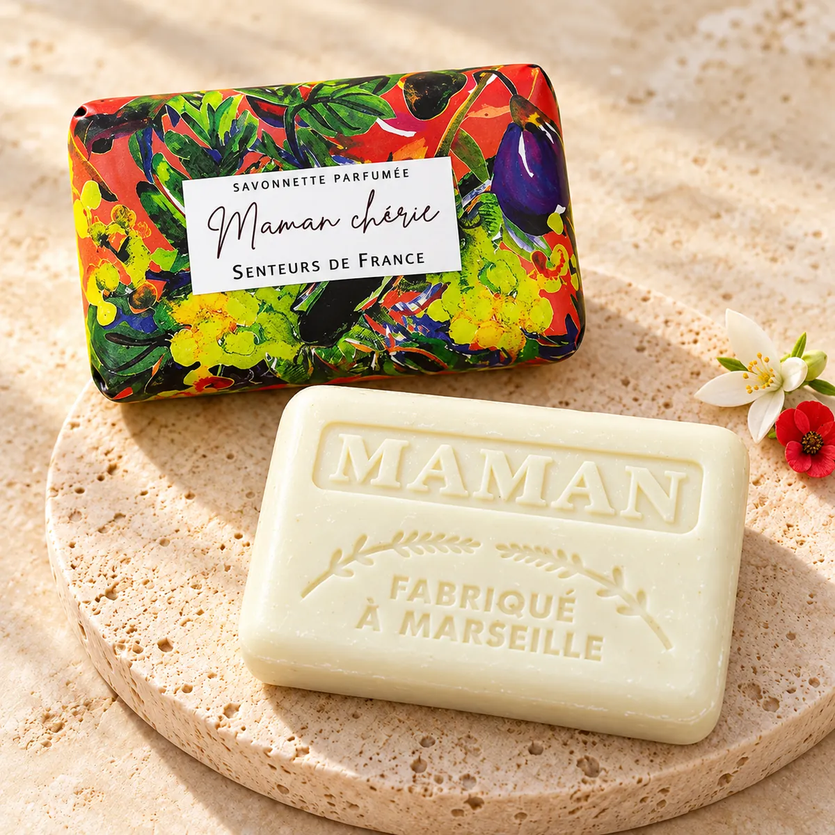 Cremefarbene Maman-chérie-Seife auf Travertin mit bunter Verpackung sowie einer weißen und einer roten Blüte.