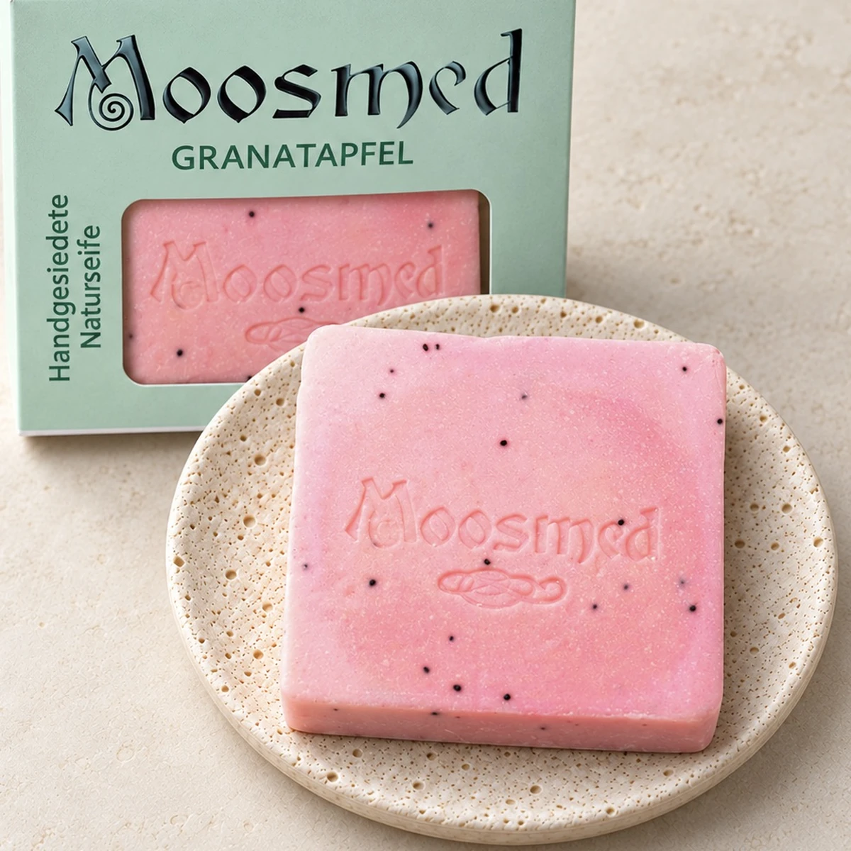 Rosafarbene Moosmed-Granatapfel-Seife mit dunklen Körnchen auf einem runden Travertinteller, dahinter die mintgrüne Verpackung.