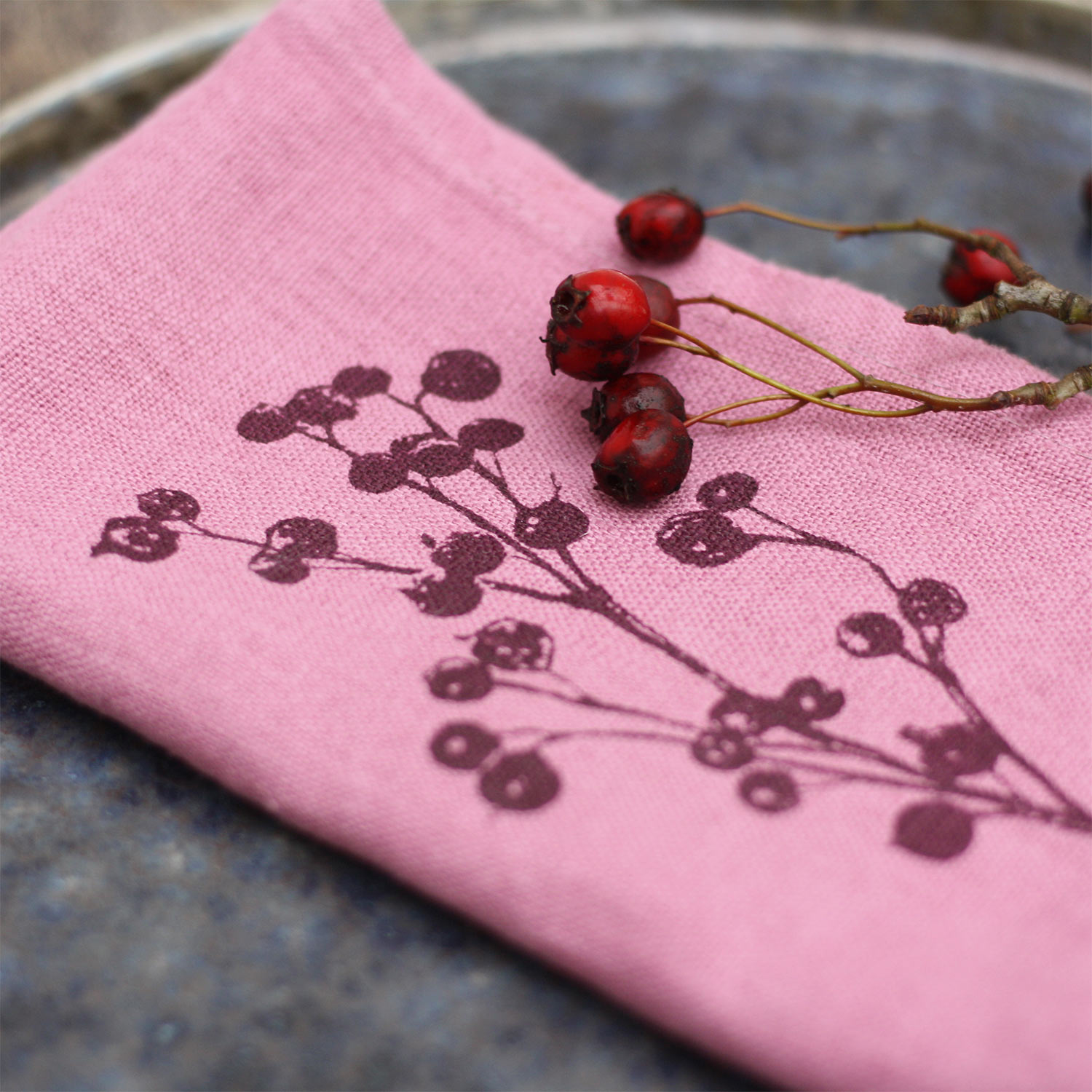 Frohstoff Leinenserviette Beerenzweig in Erika-Rosa mit feinem Beerendruck und roten Beerenzweigen auf dunklem Teller – handbedruckte Stoffserviette aus Hamburg.