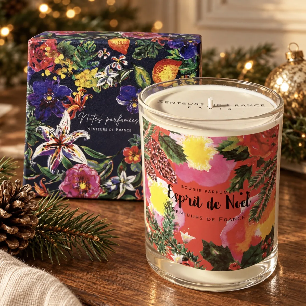 Duftkerze Esprit de Noël von Senteurs de France mit floraler Geschenkbox auf Holz, umgeben von Tannengrün, Zapfen und goldener Weihnachtsdekoration.