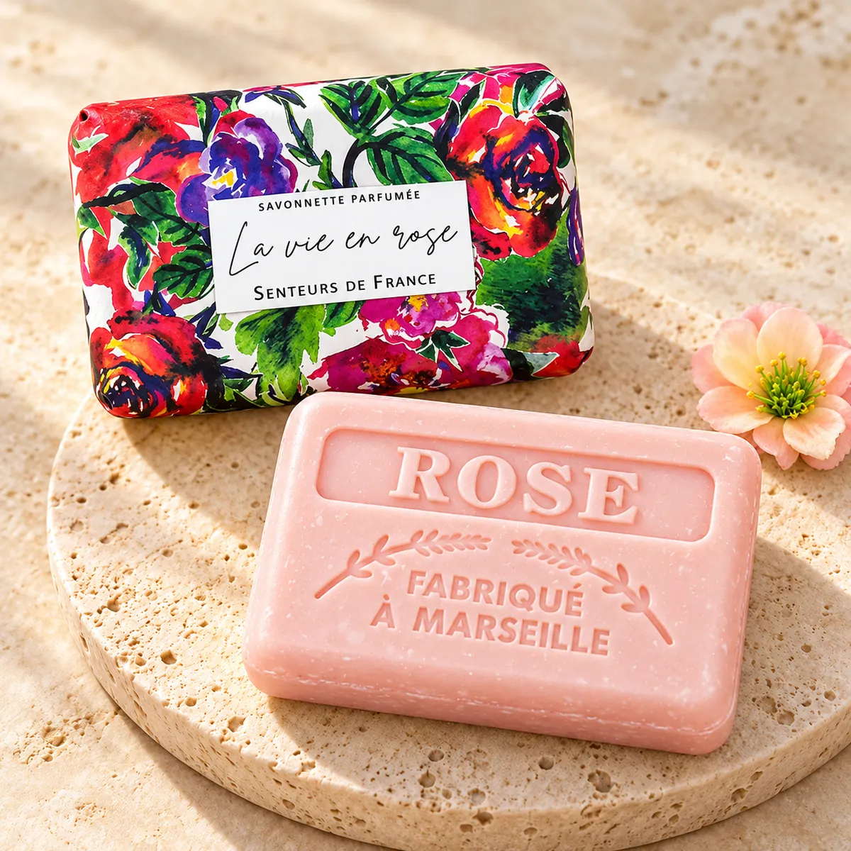 Rosafarbene La-vie-en-rose-Seife auf einem Travertinteller mit floraler Verpackung und zarter Blüte.