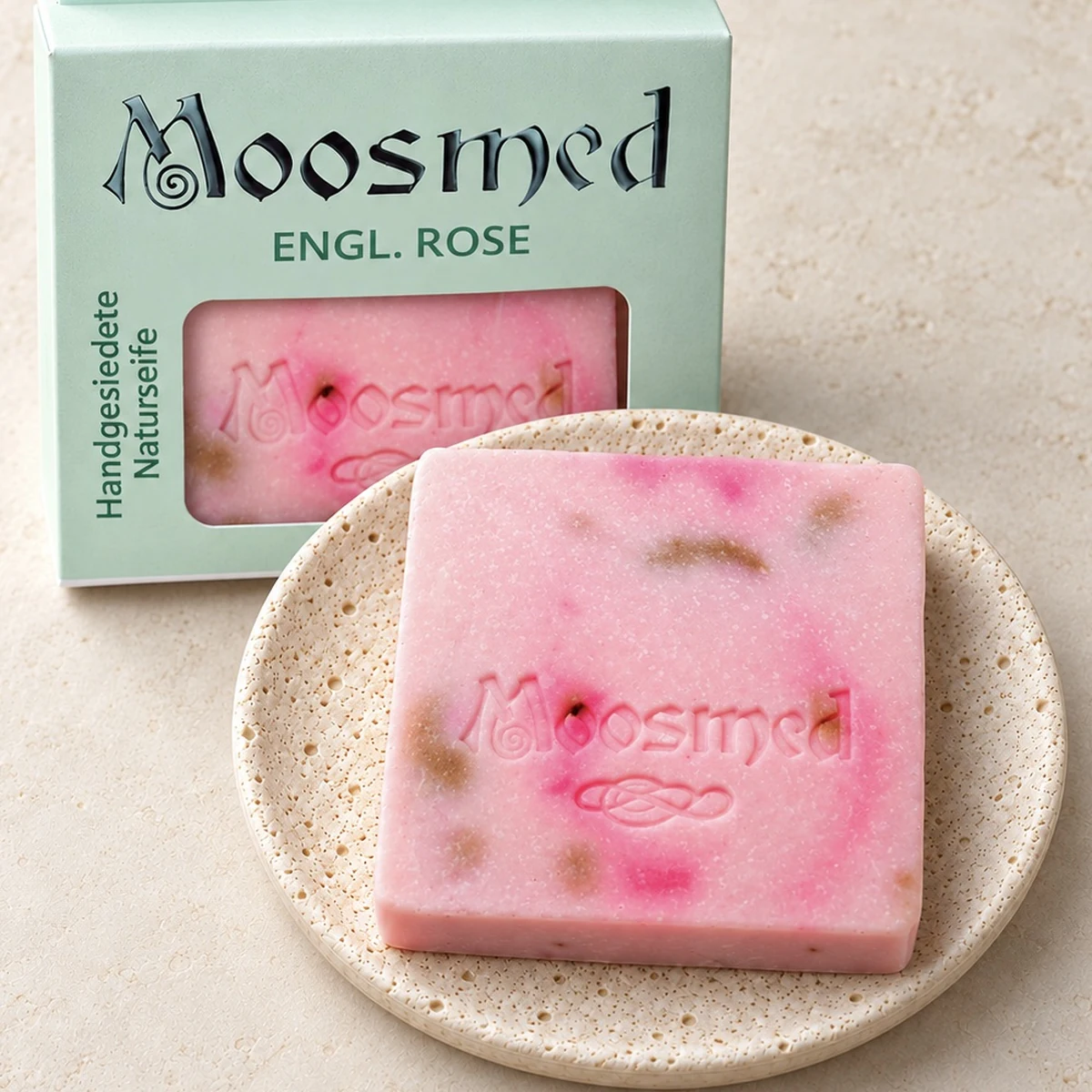 Rosafarbene Moosmed-English-Rose-Seife mit pinken und bräunlichen Akzenten auf einem runden Travertinteller, dahinter die mintgrüne Verpackung.