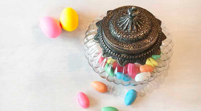 Elegante Bonbonniere aus Glas im Antiklook mit verziertem Metalldeckel, gefüllt mit bunten Zuckermandeln, auf hellem Untergrund dekorativ arrangiert.