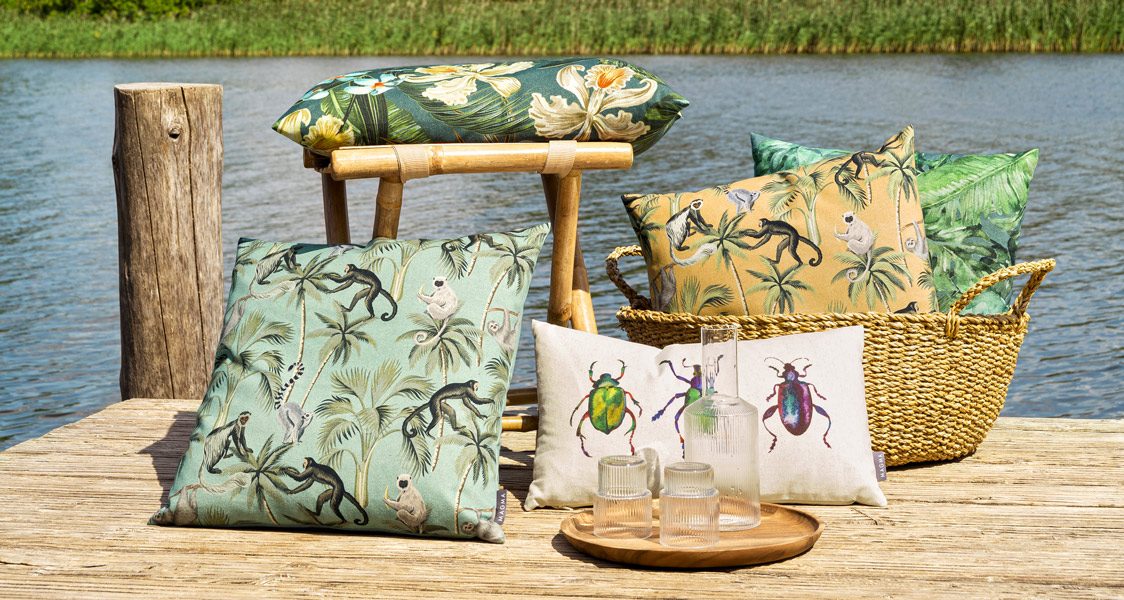 Magma Kissen „Backwoods“ mit exotischen Dschungelmotiven – Affen, Blätter und Käfer in Naturtönen, stilvoll arrangiert im sommerlichen Outdoor-Ambiente am Wasser.