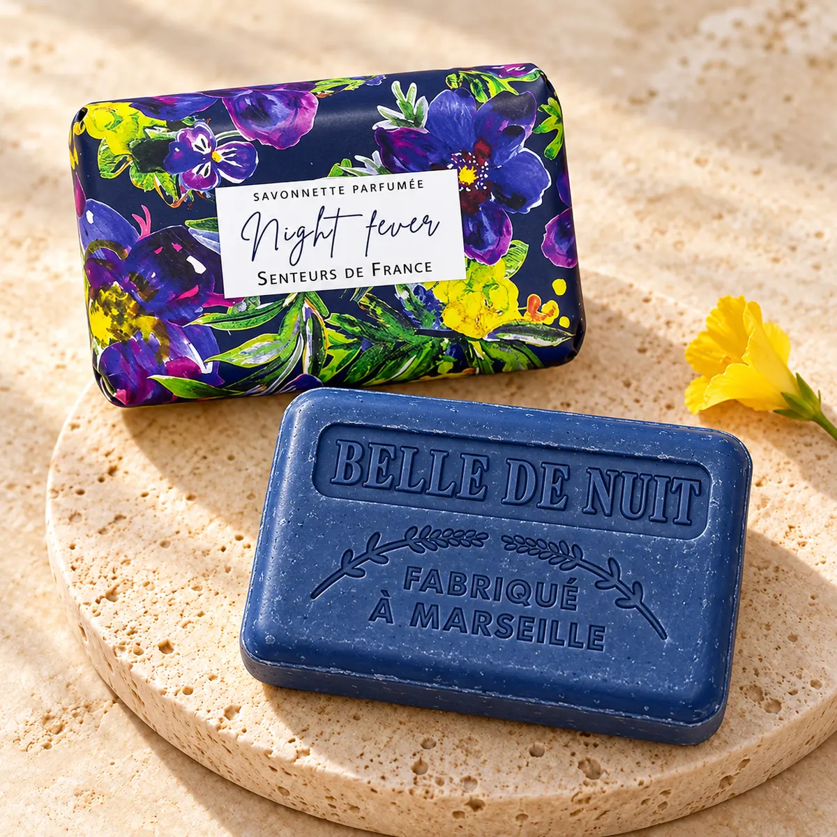 Blaue Night-fever-Seife mit Belle-de-nuit-Prägung auf Travertin, dahinter die florale Verpackung und eine gelbe Blüte.