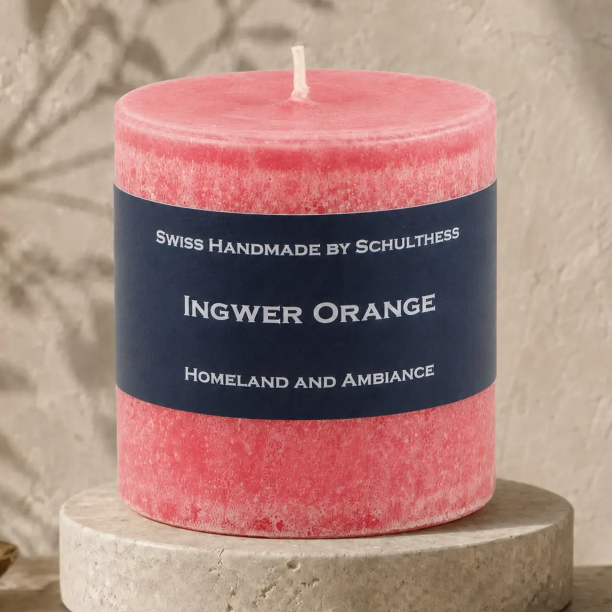 Korallrote Schulthess Duftkerze Ingwer Orange auf einem runden Travertinsockel vor einer warmen, strukturierten Wand.